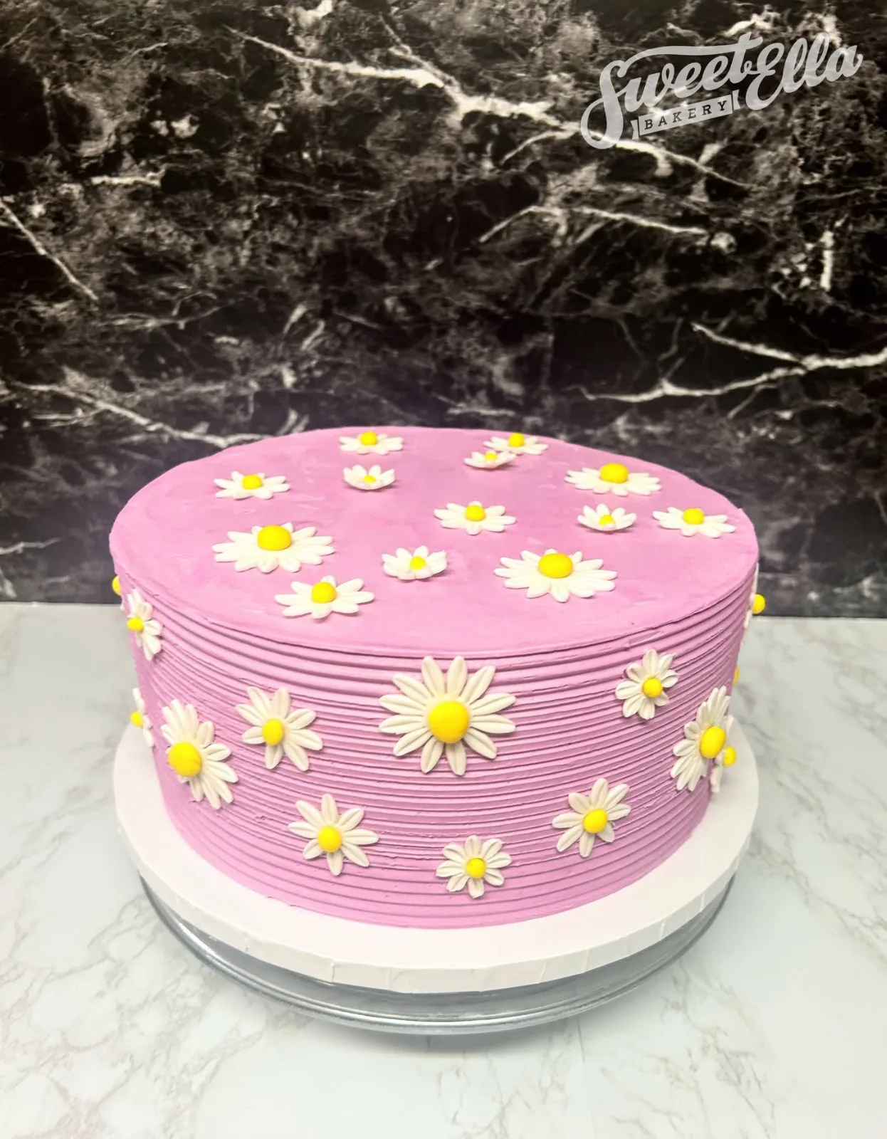 A round pink buttercream cake covered in white and yellow daisies
